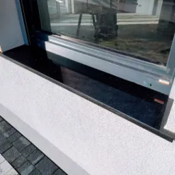 Czarny, błyszczący parapet zewnętrzny z kamienia, zamontowany pod oknem z szarą ramą, widoczny fragment elewacji budynku z szarą kostką brukową.