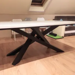 Stół z białym blatem i czarnymi, krzyżującymi się nogami w nowoczesnym wnętrzu poddasza z oknem dachowym.