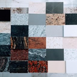 Faktoria Kamienia Grudzień Monika Juda - Zestawienie próbek różnych materiałów, w tym granitu, marmuru i konglomeratu, ułożonych na marmurowej posadzce, prezentujących bogatą paletę kolorów i tekstur.