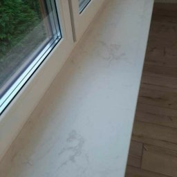 Faktoria Kamienia Grudzień Monika Juda - Jasny parapet wewnętrzny z konglomeratu z delikatnym marmurkowym wzorem, zamontowany w białym oknie, widok na zieleń za oknem, obok widoczna drewniana podłoga.