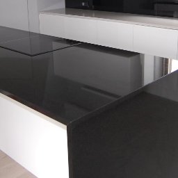 Faktoria Kamienia Grudzień Monika Juda - Czarny, błyszczący blat z kamienia z wbudowaną płytą grzewczą, zamontowany w nowoczesnej kuchni w Alwerni. Minimalistyczny design z białymi szafkami w tle.