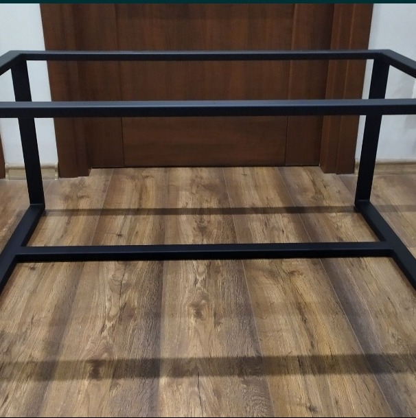 Metalowa rama stołu z profili kwadratowych, pomalowana na czarno, ustawiona na podłodze z paneli imitujących drewno, na tle zamkniętych drzwi.