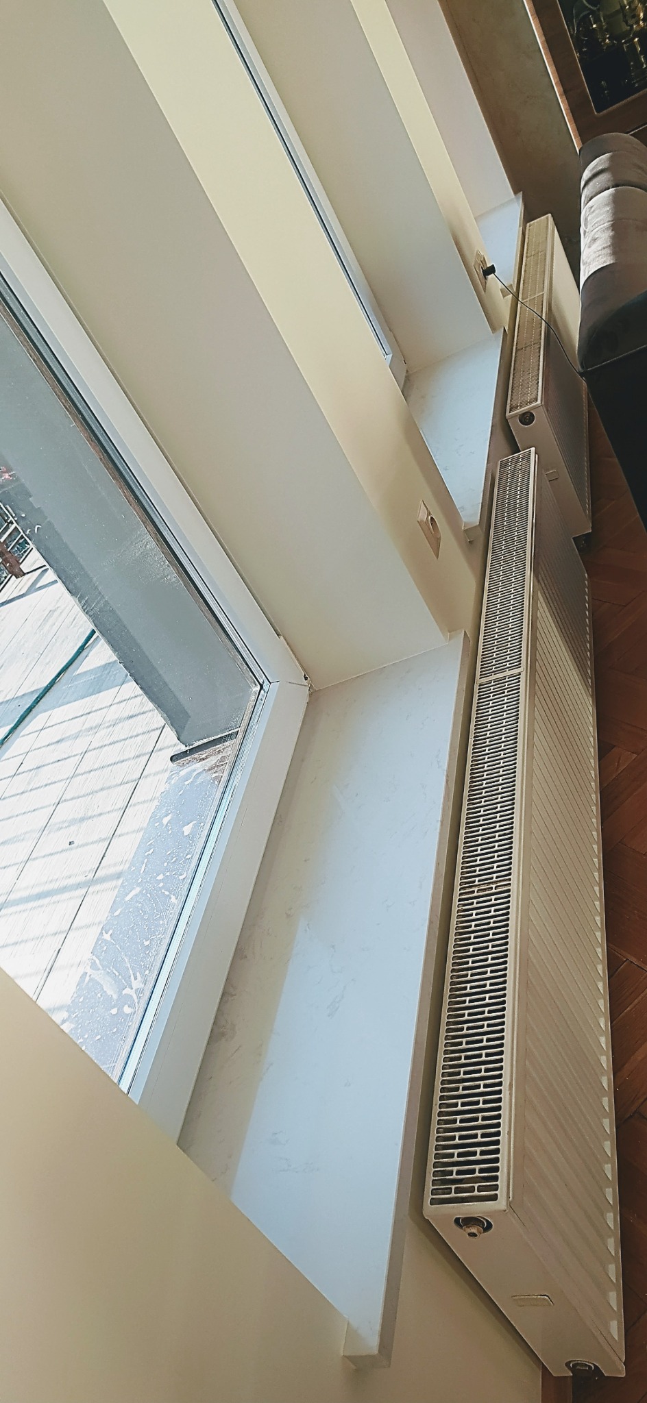 Białe grzejniki żeberkowe zamontowane pod oknem w jasnym pomieszczeniu z drewnianą podłogą, widoczne ślady czyszczenia okna.