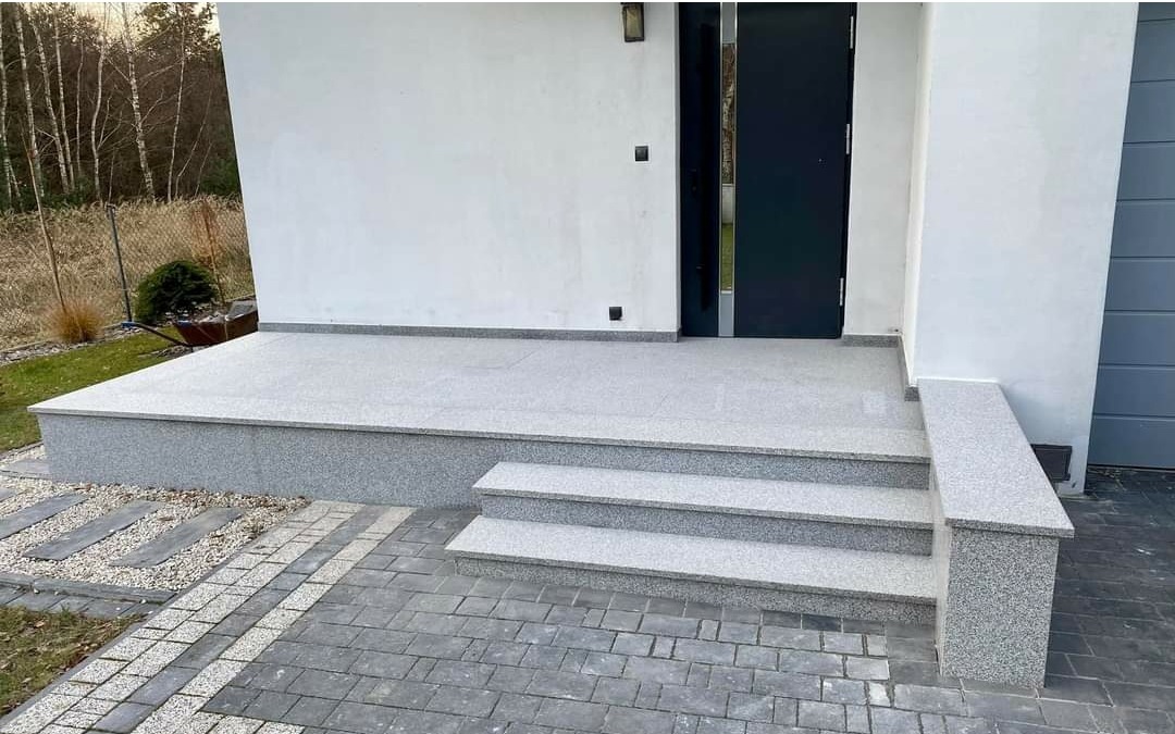 Nowoczesne wejście do domu z szarego granitu: trzystopniowe schody, podest i ścieżka z kostki brukowej, minimalistyczna elewacja z czarnymi drzwiami.