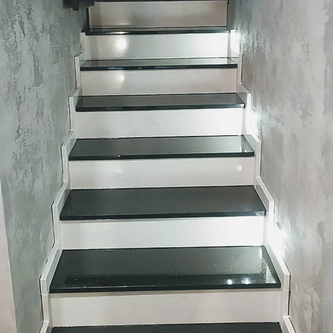 Nowoczesne schody z czarnymi stopniami i białymi podstopnicami, oświetlenie punktowe przy ścianie z dekoracyjnym tynkiem.