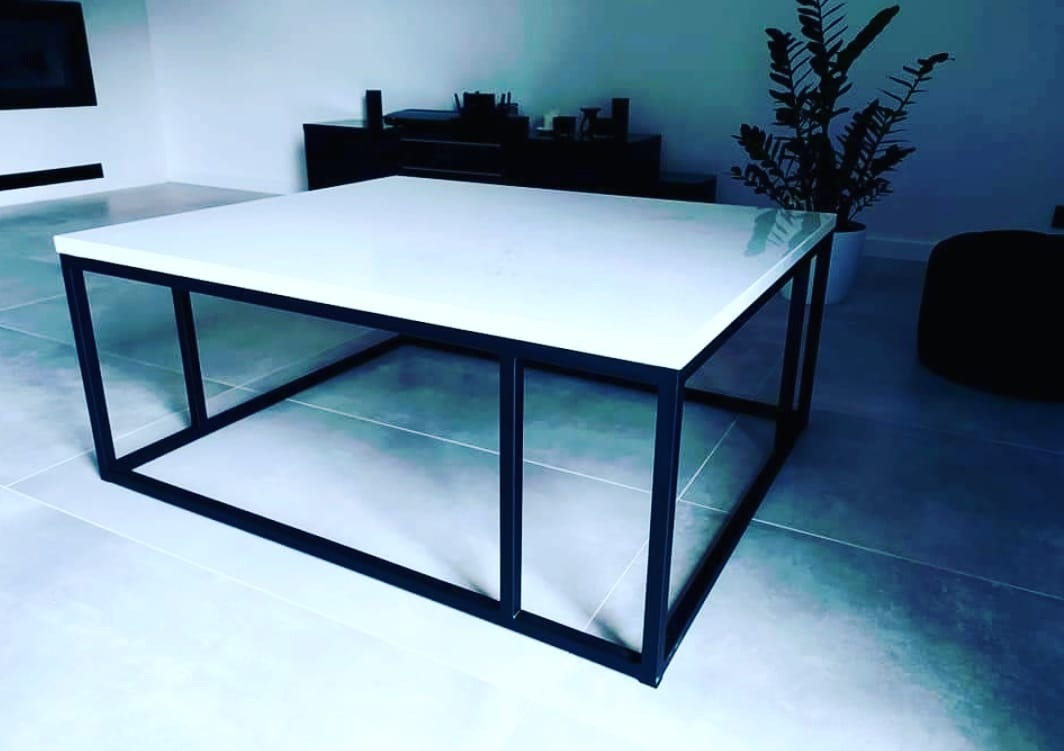 Nowoczesny stolik kawowy z białym blatem przypominającym konglomerat i czarną, stalową ramą, ustawiony na szarych płytkach podłogowych w minimalistycznym salonie.