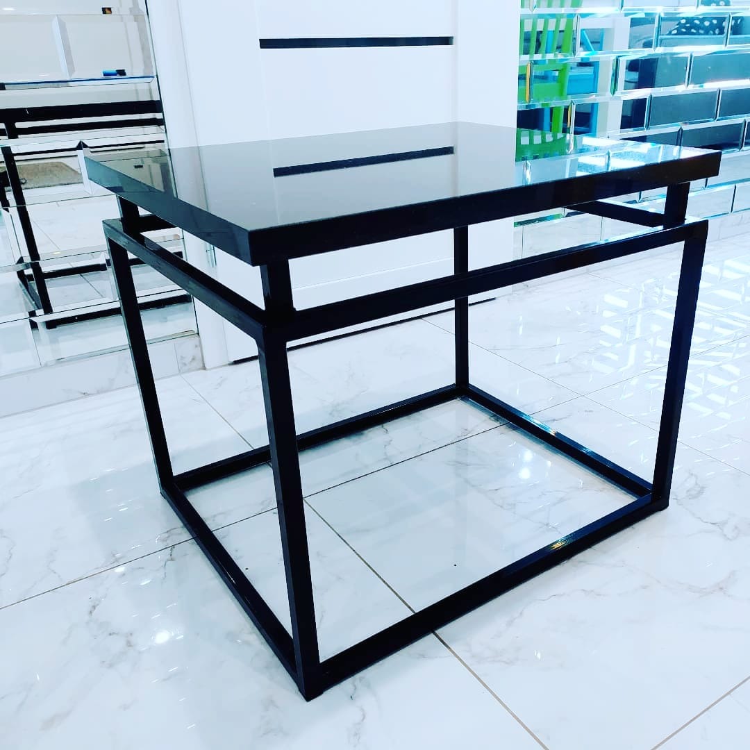 Stolik kawowy z czarnym, lustrzanym blatem i metalową, geometryczną podstawą w stylu industrialnym, na białej marmurowej podłodze, odbijający światło w minimalistycznym wnętrzu.