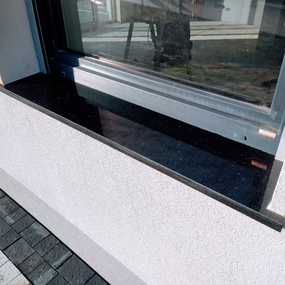 Czarny, błyszczący parapet zewnętrzny z kamienia, zamontowany pod oknem z szarą ramą, widoczny fragment elewacji budynku z szarą kostką brukową.