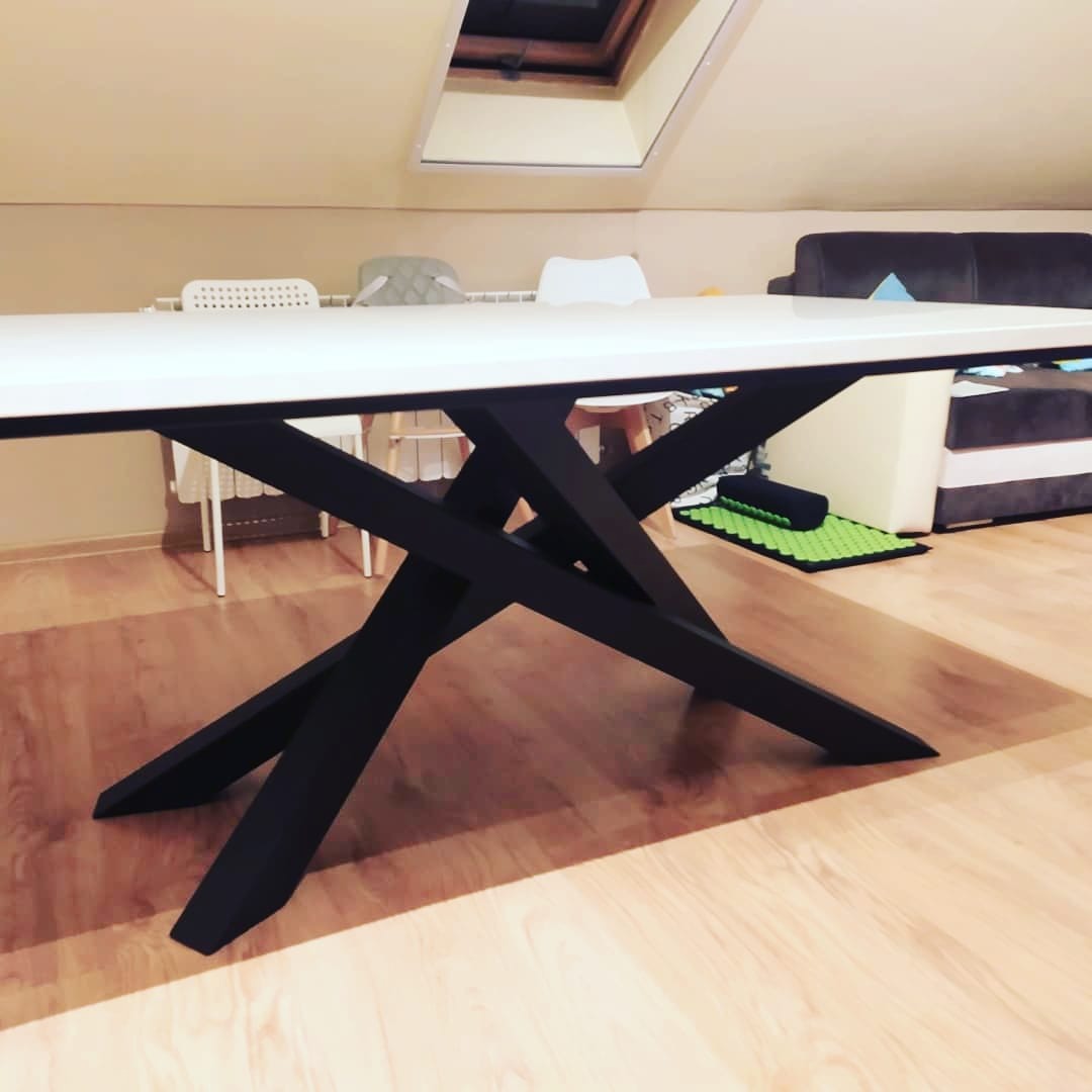 Stół z białym blatem i czarną, geometryczną podstawą w nowoczesnym wnętrzu z drewnianą podłogą i oknem dachowym.