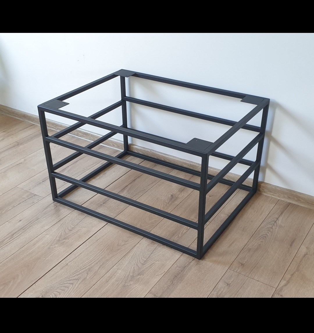 Czarna, spawana konstrukcja stalowa w kształcie sześcianu, z trzema poziomami, umieszczona na podłodze z paneli, na tle białej ściany, z widocznymi spawami w narożnikach.