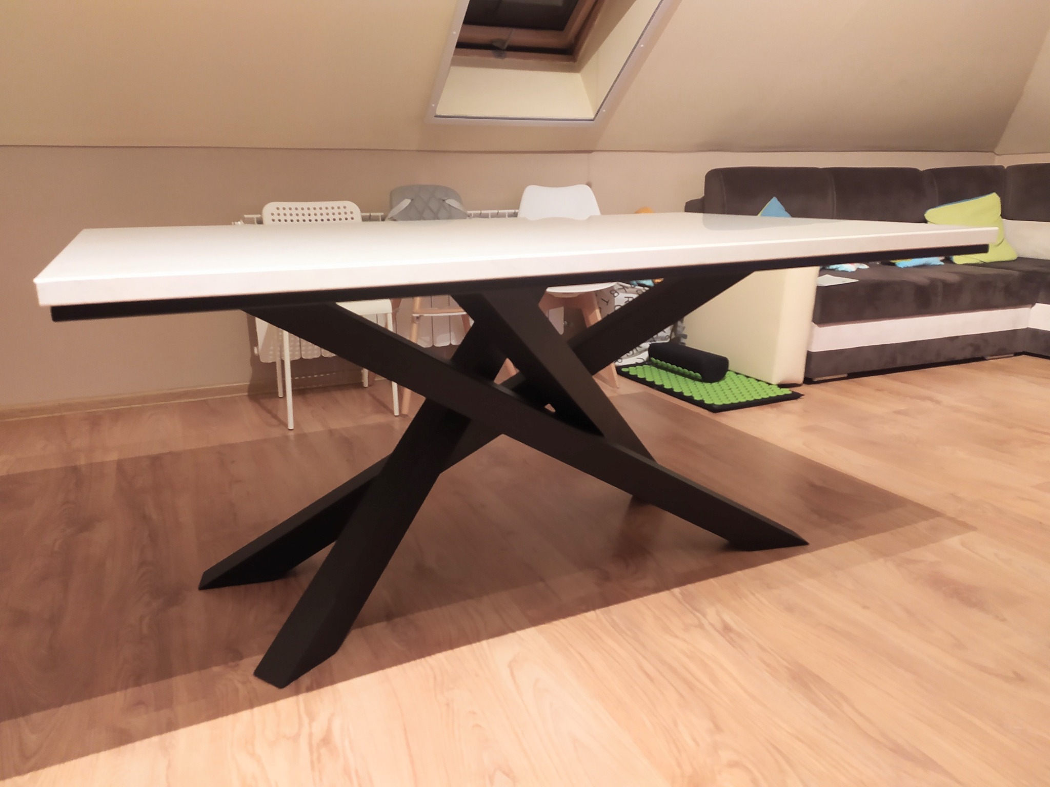 Stół z białym blatem i czarnymi, krzyżującymi się nogami w nowoczesnym wnętrzu poddasza z oknem dachowym.