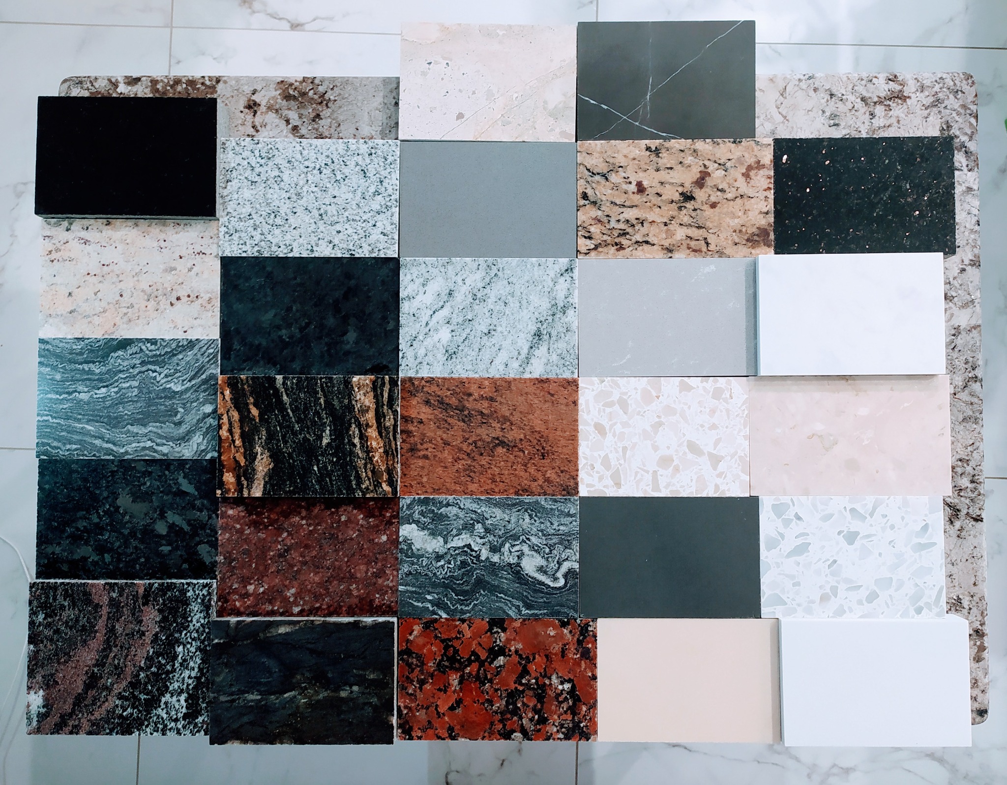 Zestawienie próbek różnych materiałów, w tym granitu, marmuru i konglomeratu, ułożonych na marmurowej posadzce, prezentujących bogatą paletę kolorów i tekstur.
