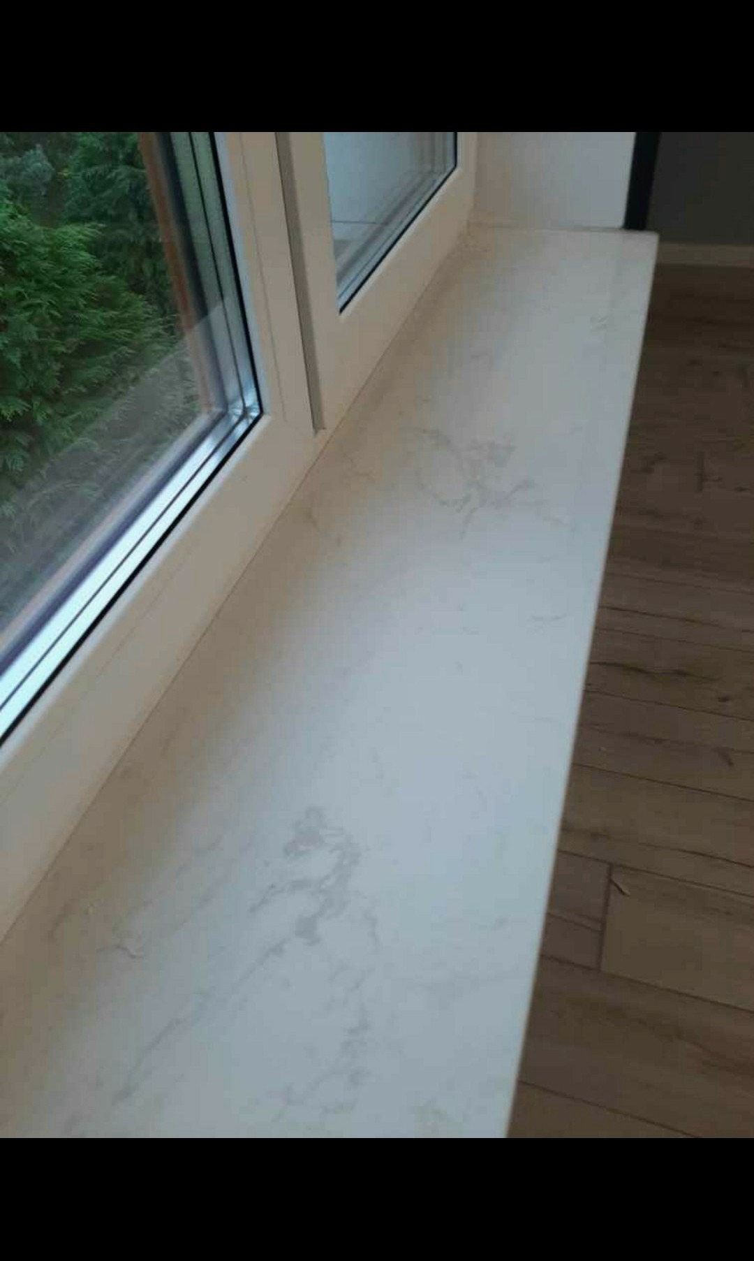 Jasny parapet wewnętrzny z konglomeratu z delikatnym marmurkowym wzorem, zamontowany w białym oknie, widok na zieleń za oknem, obok widoczna drewniana podłoga.