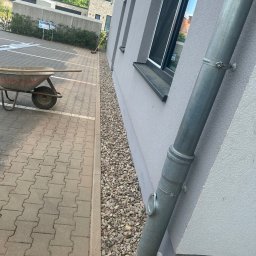 Patryk Wieczorek - Szara, teksturowana elewacja nowoczesnego budynku z metalową rynną spustową i oknem. Obok schludna opaska żwirowa i chodnik z kostki brukowej; w tle taczka.