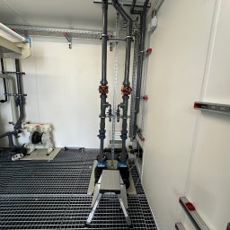 Patryk Wieczorek - Szczegółowa instalacja szarych rur z pomarańczowymi i niebieskimi zaworami, zamontowana na białych ścianach w pomieszczeniu technicznym z metalową kratą podłogową. Widoczna pompa ARO i mały podest...
