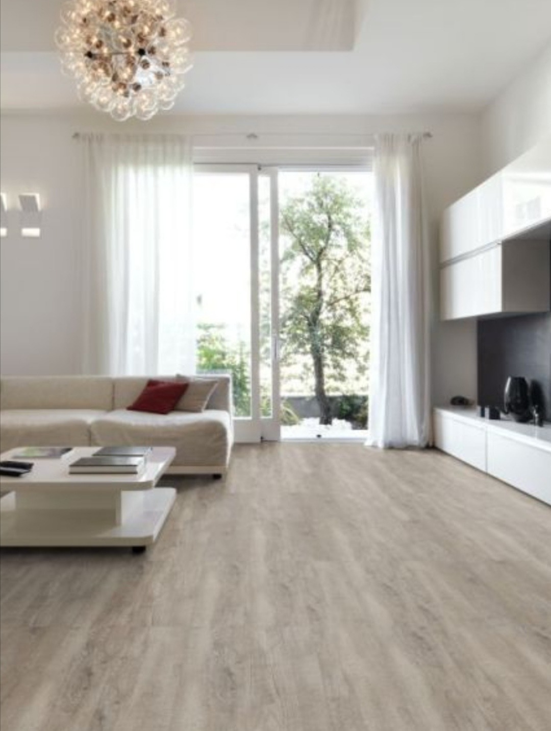 Jasne panele podłogowe w salonie z białymi ścianami, sofą, stolikiem kawowym i dużym oknem balkonowym z widokiem na zieleń.