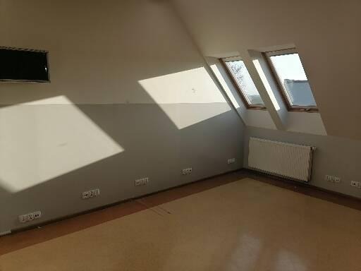 Poddasze z trzema oknami dachowymi, pomalowane na szaro ściany, widoczne gniazdka elektryczne przy podłodze, grzejnik pod oknami, drewniana podłoga.