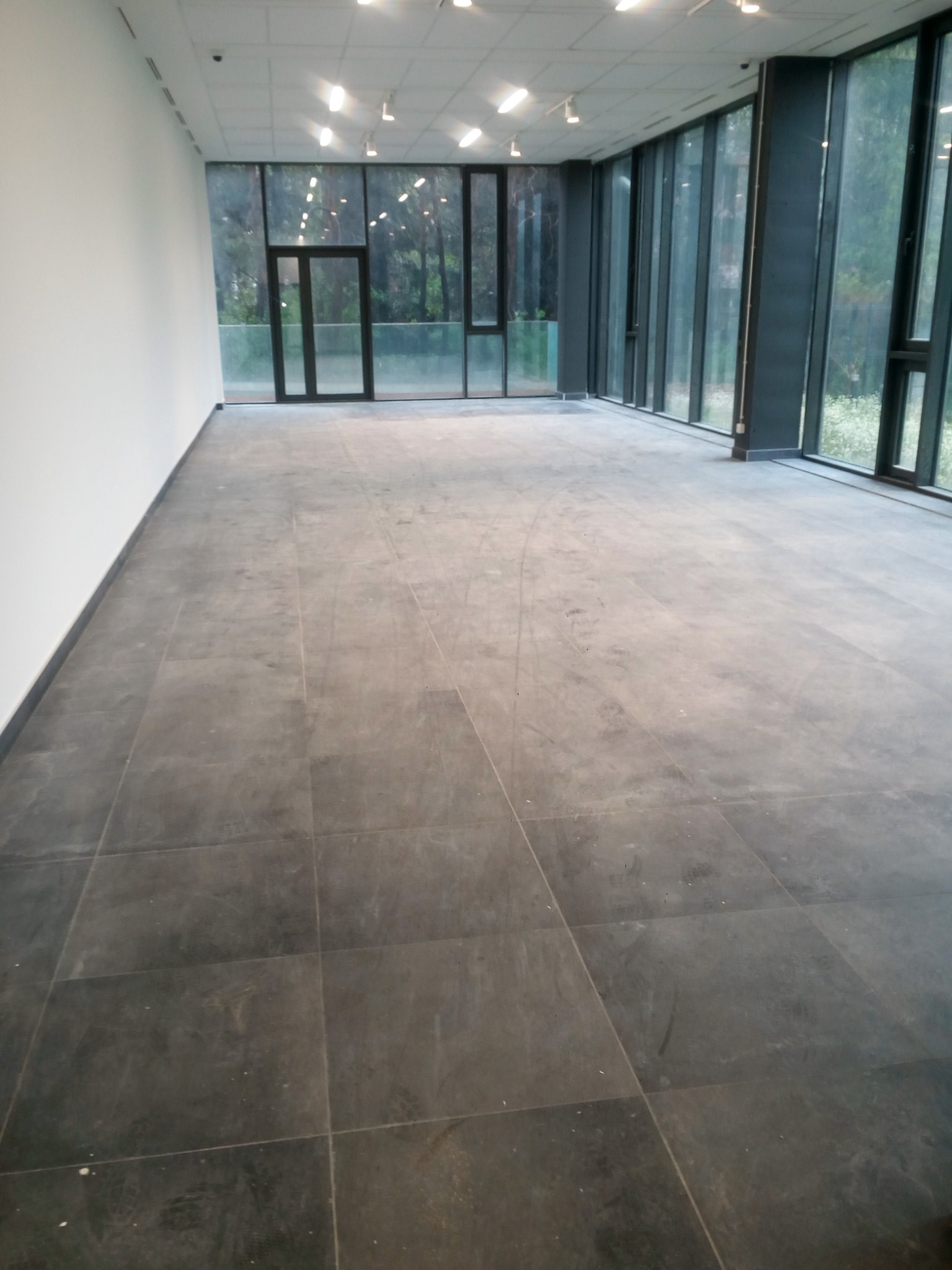 Przestronne, puste wnętrze biurowe z szarymi płytkami podłogowymi, dużymi oknami od podłogi do sufitu i białymi ścianami, oświetlone lampami sufitowymi.