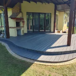 Szara dwupoziomowa taras kompozytowy z zaokrąglonymi krawędziami, wykończona kostką brukową, przylegająca do domu z ceglanym grillem i dużymi oknami, otoczona trawnikiem.