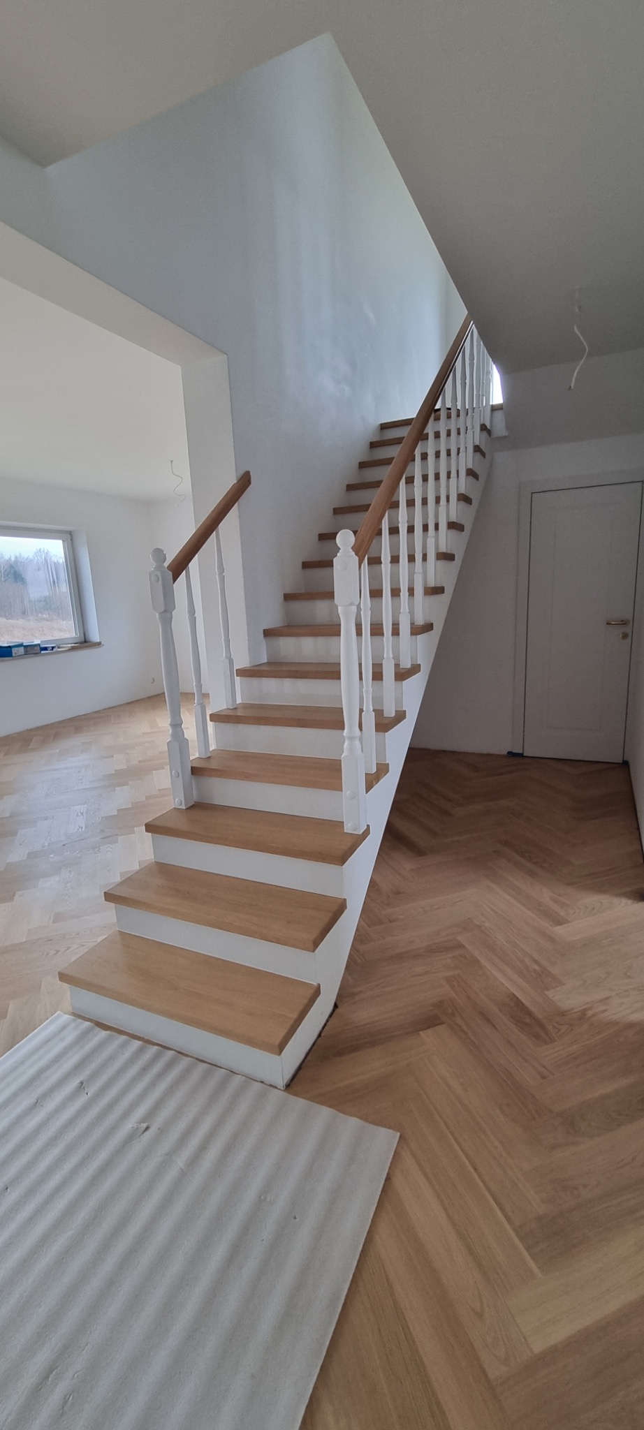 Jasne drewniane schody z białymi balustradami i podstopnicami, parkiet ułożony w jodełkę, widok z dołu schodów w nowym domu.