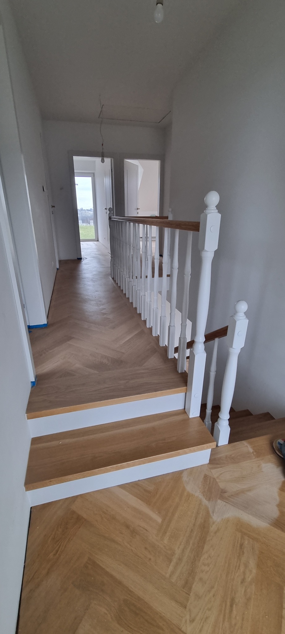Korytarz z drewnianą podłogą ułożoną w jodełkę i białymi schodami z drewnianą poręczą. Widoczne niedoskonałości na podłodze sugerujące prace wykończeniowe.