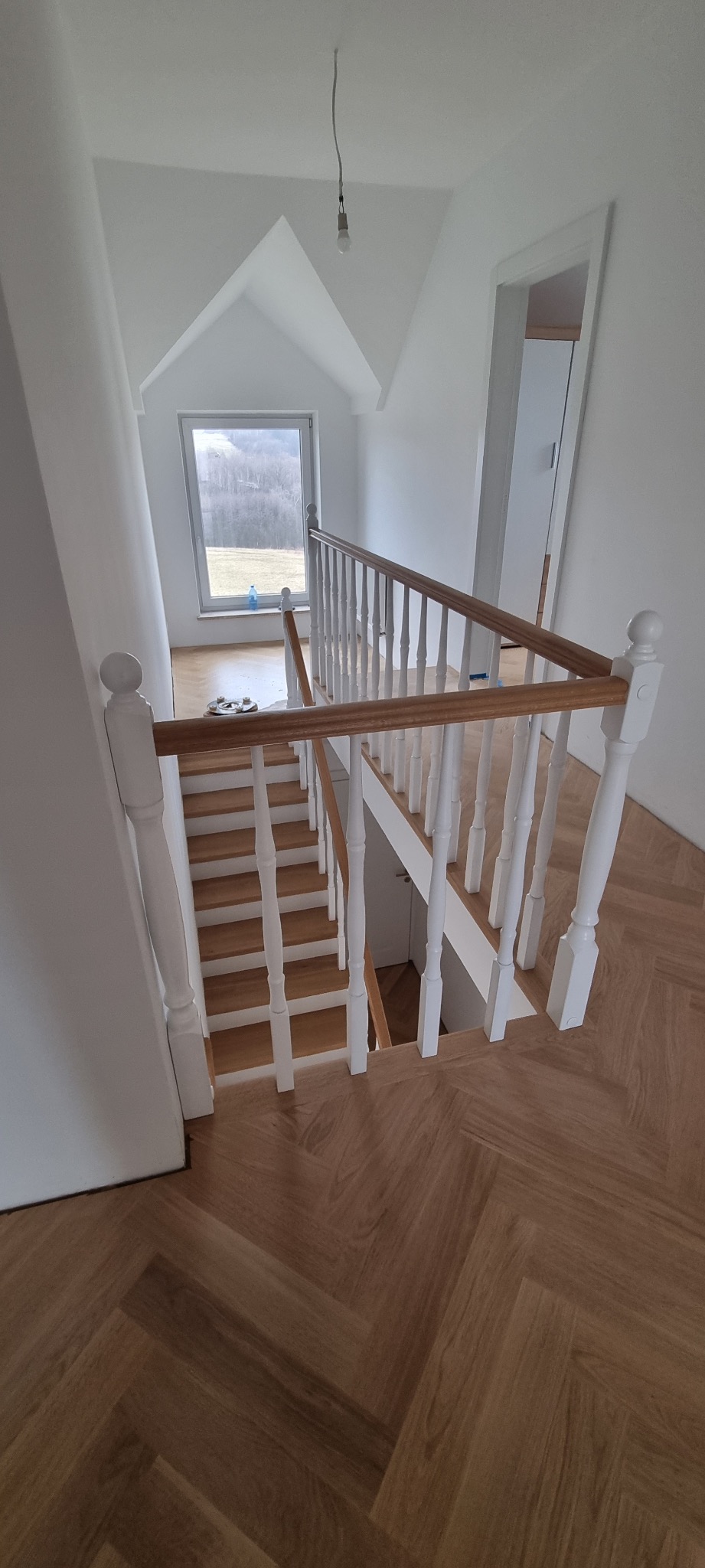 Jasne drewniane schody z białymi balustradami w nowym domu, parkiet ułożony w jodełkę, widok na okno z zielenią w tle.