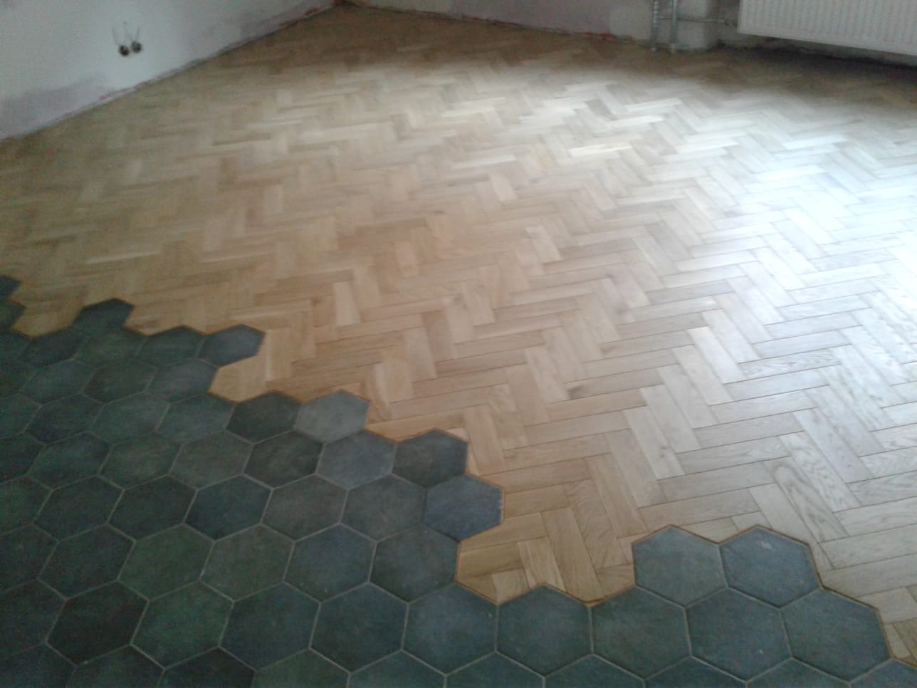 Fragment podłogi w trakcie remontu: klasyczny parkiet ułożony we wzór jodełki sąsiaduje z ciemnymi, heksagonalnymi płytkami podłogowymi, widoczne niedokończone wykończenie przy ścianie.