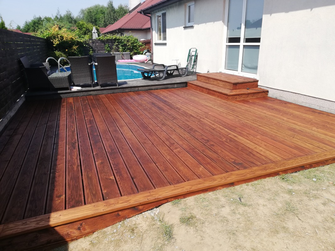 Nowoczesny taras z ciemnego drewna z niskimi schodkami, przylegający do białego domu z basenem i meblami ogrodowymi w tle.