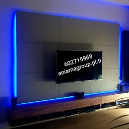 Ściana TV wykończona szarymi panelami z niebieskim podświetleniem LED, drewniana szafka RTV z widocznym sprzętem audio-video.