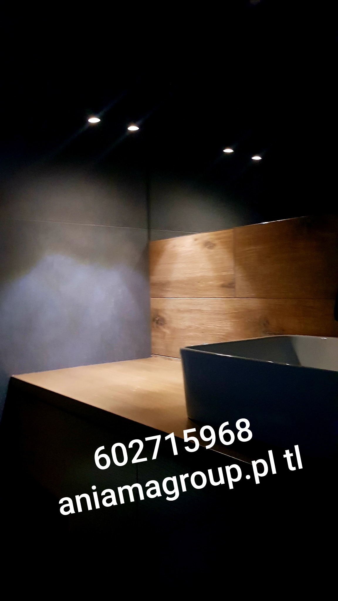 Nowoczesna łazienka z szarą ścianą z gładzią, drewnianą okładziną i minimalistyczną, ciemną umywalką nablatową podświetlona punktowymi lampami sufitowymi.