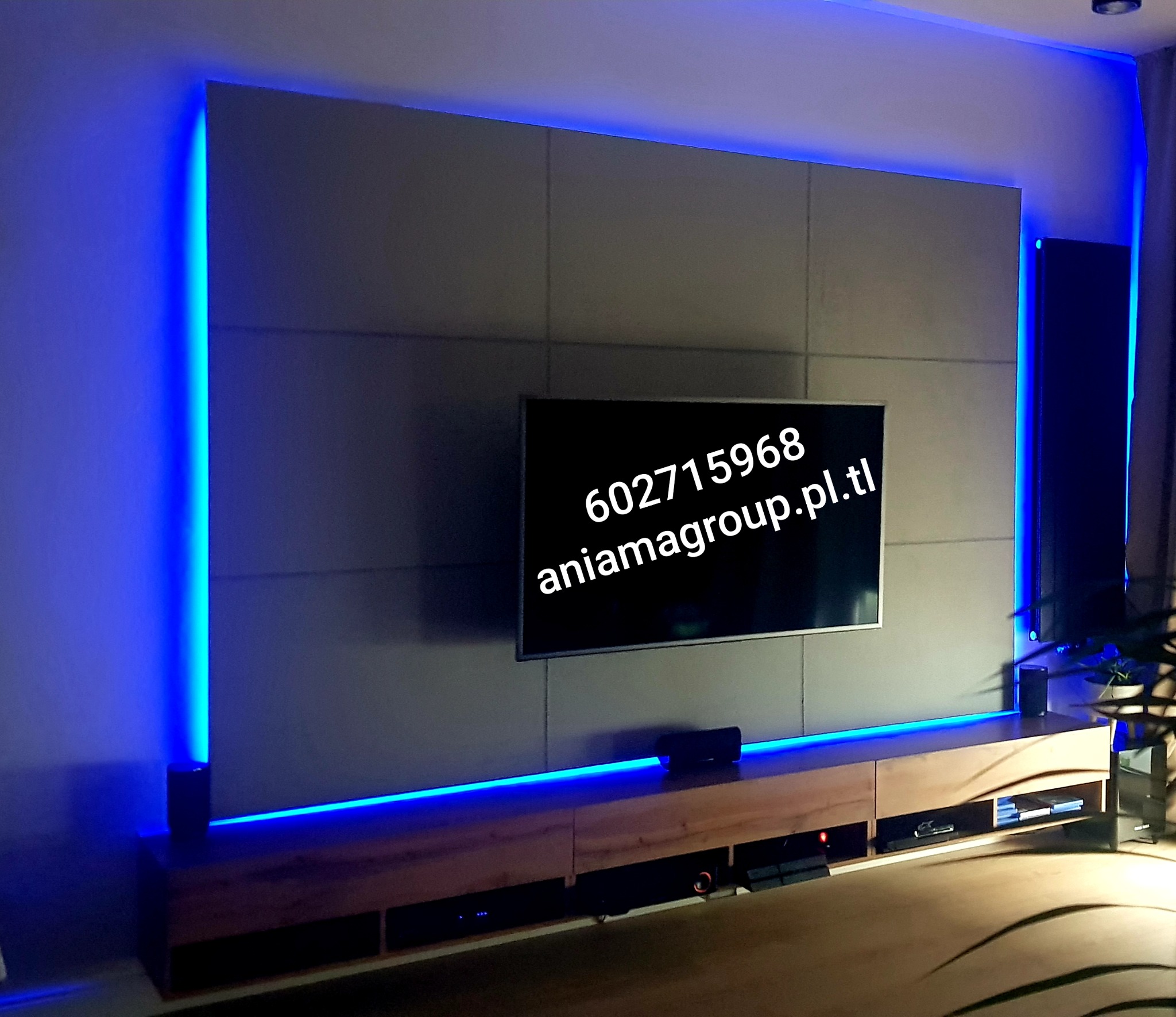 Ściana TV wykończona szarymi panelami z niebieskim podświetleniem LED, drewniana szafka RTV z widocznym sprzętem audio-video.