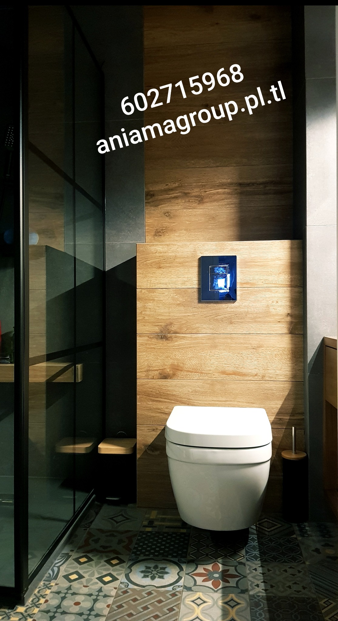 Nowoczesna toaleta wisząca na tle ściany wyłożonej panelami imitującymi drewno, z widoczną szklaną kabiną prysznicową i wzorzystą podłogą w łazience.