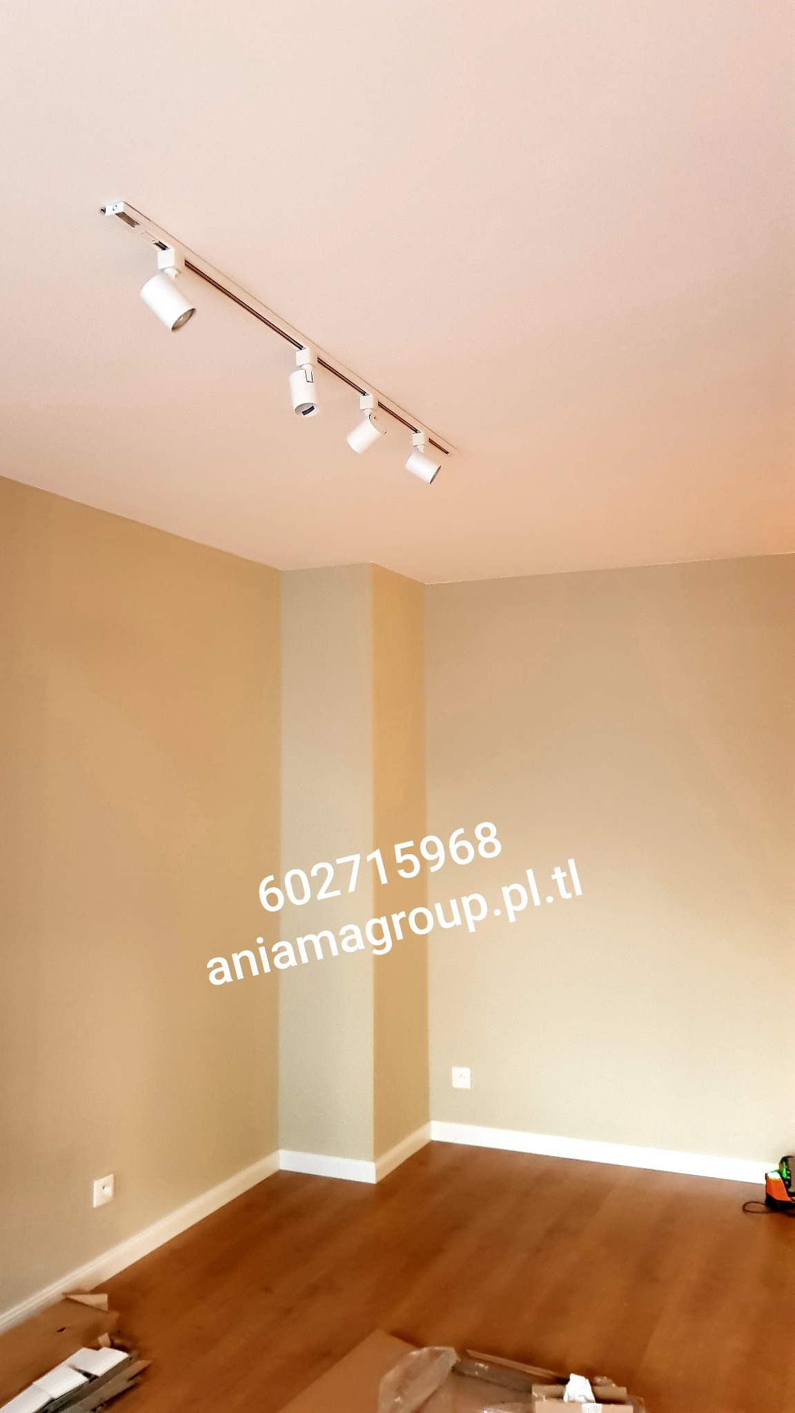 Beżowe ściany w pustym pokoju z drewnianą podłogą, biała listwa przypodłogowa i reflektory sufitowe na szynie; widoczny fragment podłogi z kartonami.