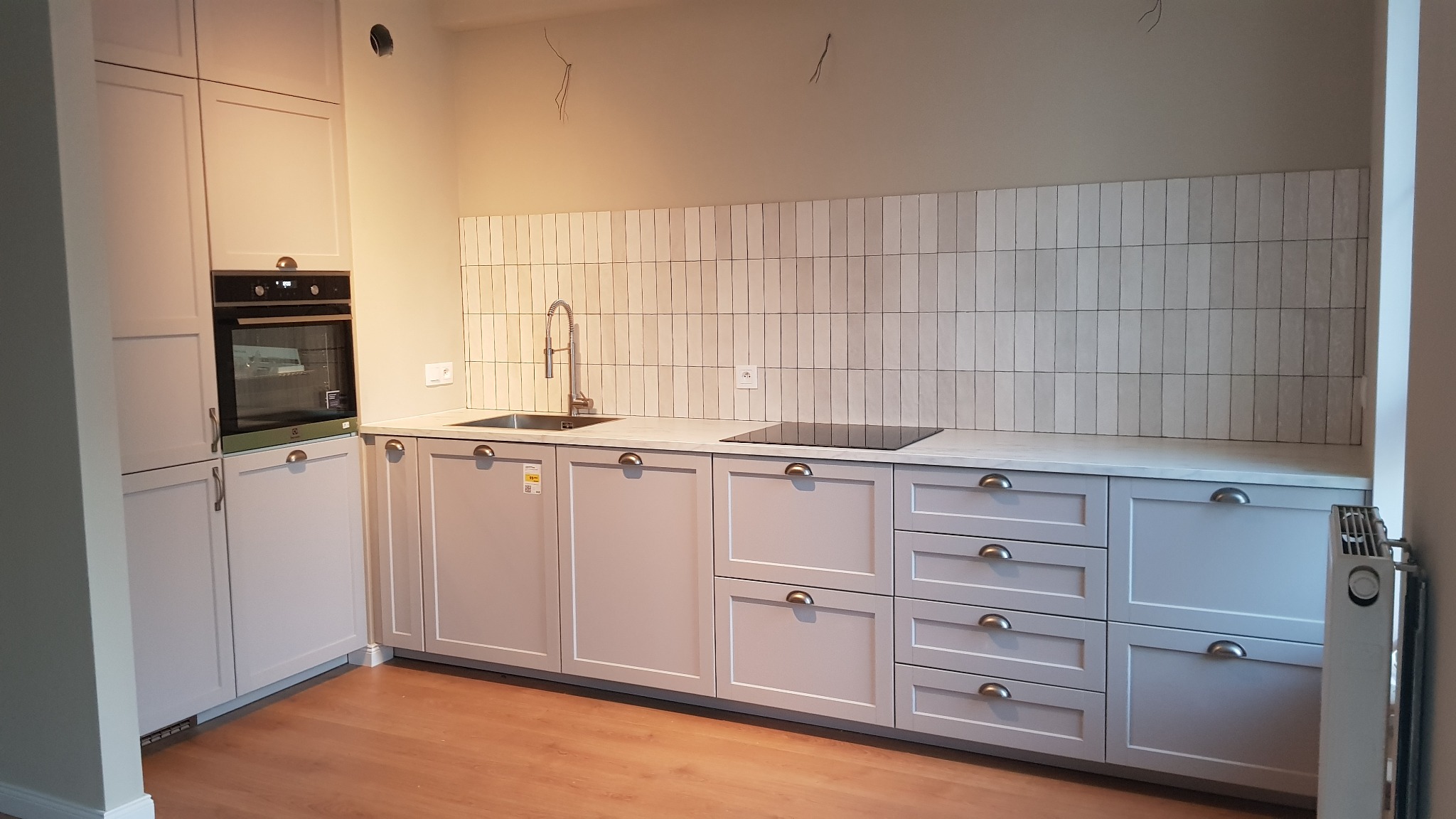 Jasna kuchnia z szarymi szafkami, białym blatem i pionowymi płytkami na ścianie, widoczny zlew, płyta indukcyjna i piekarnik.