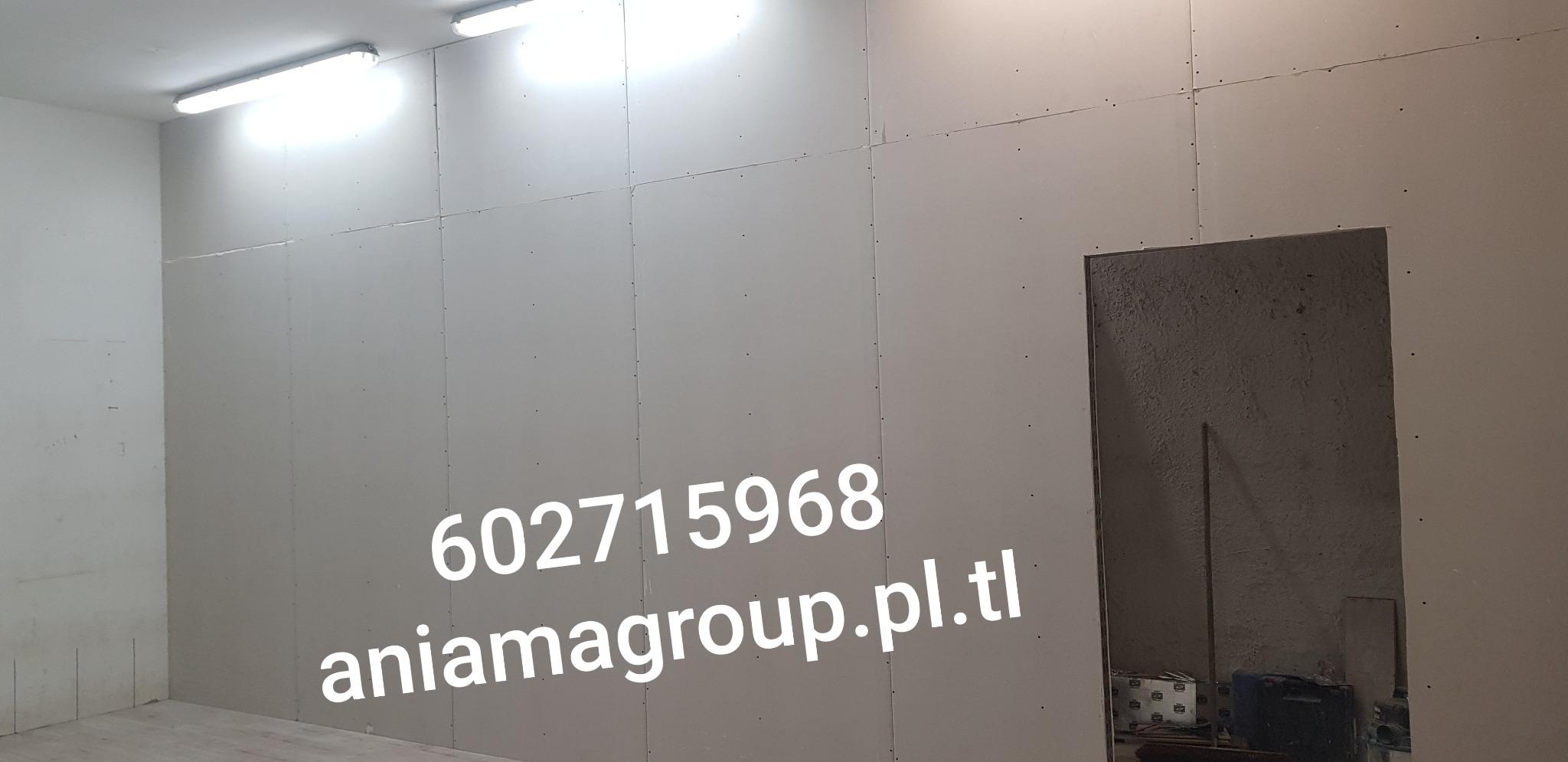 Ściana z płyt kartonowo-gipsowych w trakcie montażu, widoczne łączenia płyt, oświetlenie sufitowe oraz otwór drzwiowy z surowym wykończeniem.