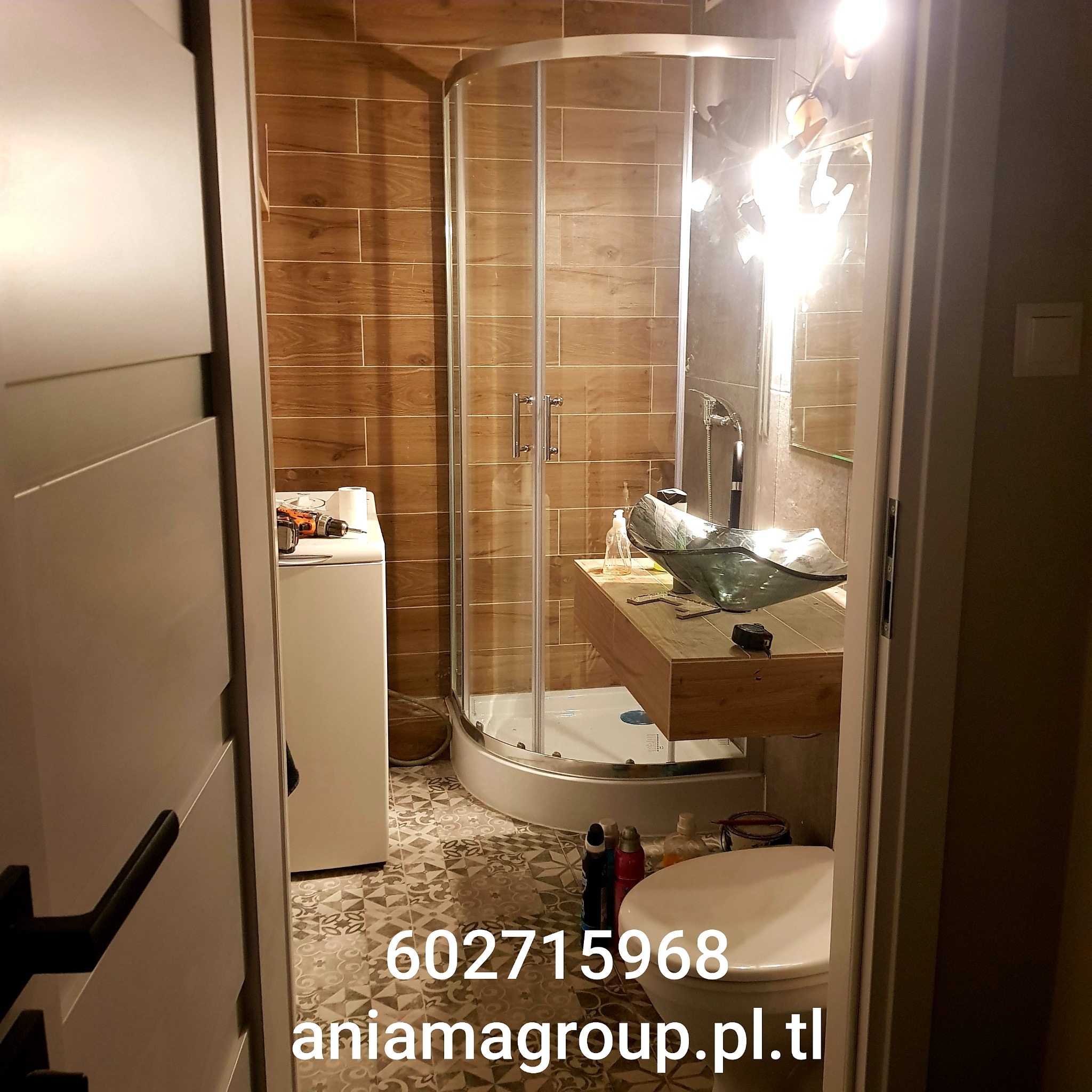 Ukończona łazienka z kabiną prysznicową, umywalką na drewnianym blacie, wzorzystą podłogą i pralką z wiertarką, widok zza uchylonych drzwi.