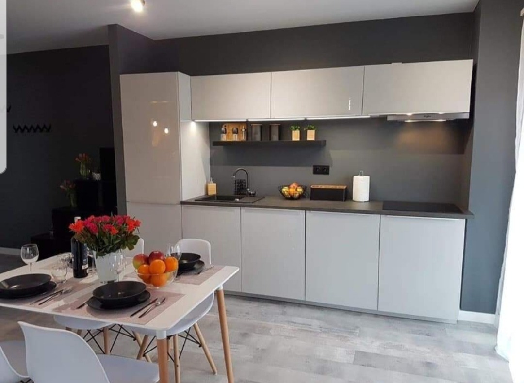 Nowoczesna kuchnia w odcieniach szarości z białymi frontami szafek, czarnym blatem i drewnianymi akcentami na krzesłach przy stole z zastawą i owocami.