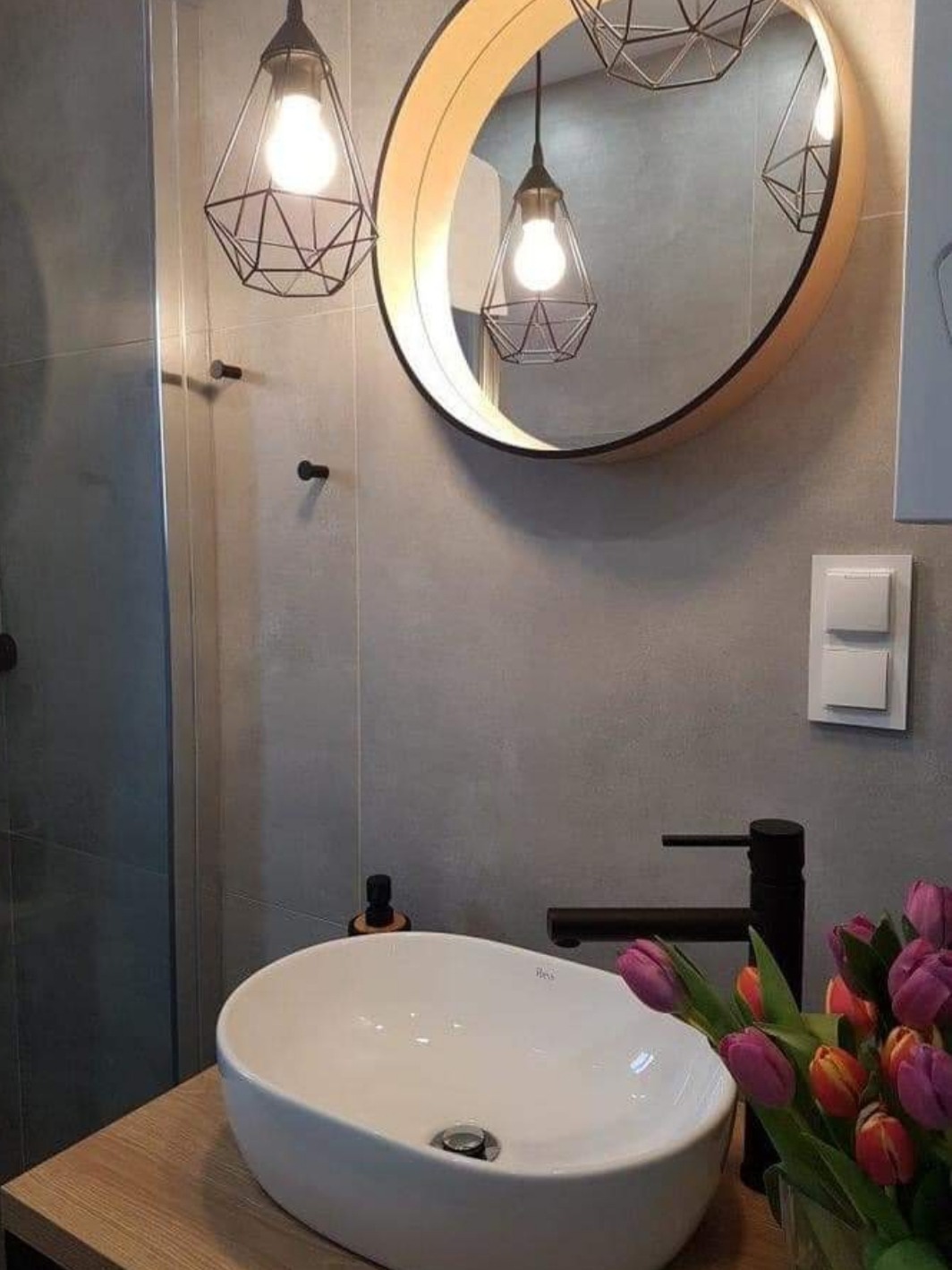 Umywalka nablatowa w nowoczesnej łazience z szarymi ścianami, okrągłym lustrem z podświetleniem LED, czarną armaturą i bukietem tulipanów.