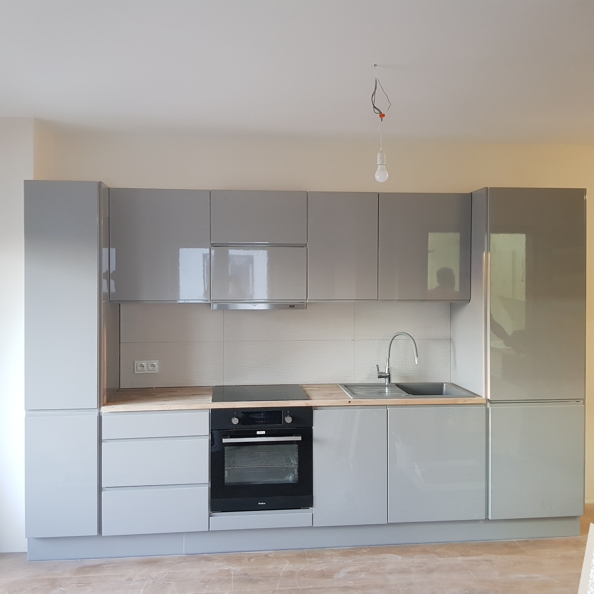 Nowoczesna, szara zabudowa kuchenna z drewnianym blatem i czarnym piekarnikiem marki Amica, z niedokończonym oświetleniem sufitowym.