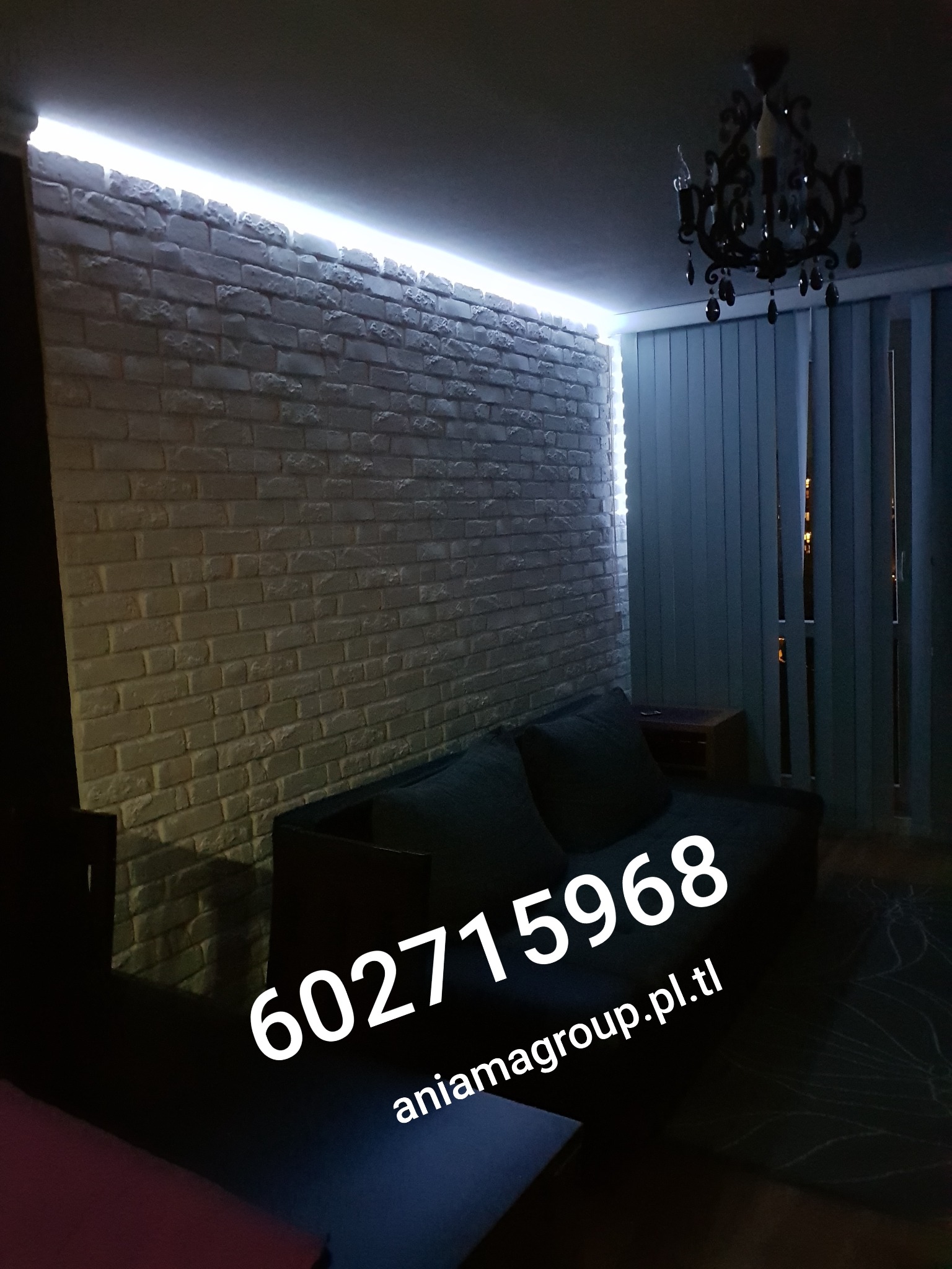 Ściana z białej cegły dekoracyjnej w salonie, oświetlona listwą LED pod sufitem, z widocznym fragmentem sofy i żyrandolem w stylu retro.