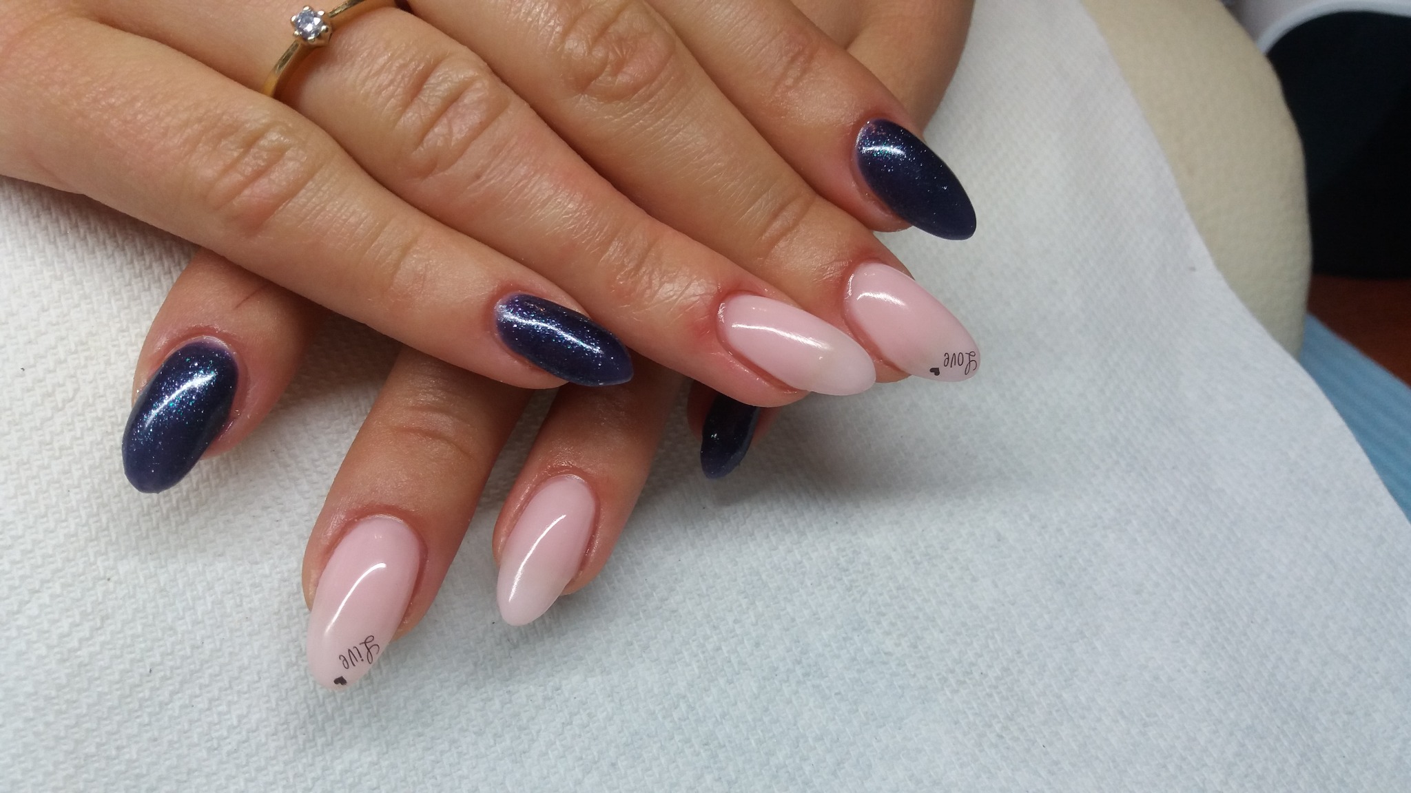 Dłonie z manicure hybrydowym: dwa paznokcie w kolorze głębokiego granatu z brokatem, trzy w odcieniu jasnego różu, jeden z napisem 'love' i małym serduszkiem, pierścionek na palcu wskazującym.