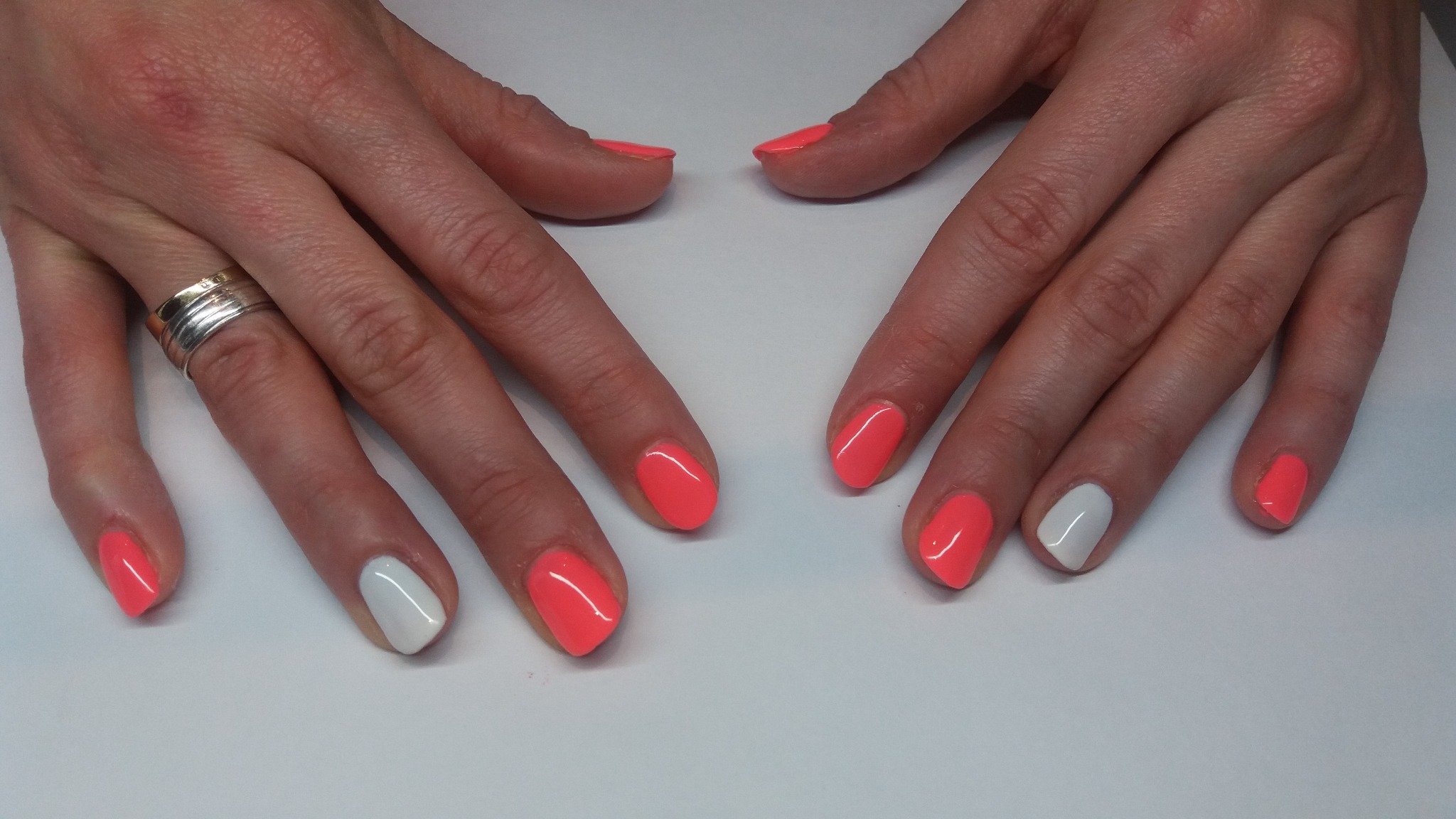 Dłonie z pomalowanymi paznokciami: koralowy lakier na większości paznokci, z wyjątkiem dwóch pomalowanych na biało, na palcu serdecznym prawej dłoni widoczny srebrny pierścionek.