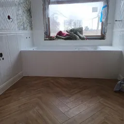 Wnętrze łazienki w trakcie prac wykończeniowych: ułożona podłoga z płytek imitujących drewno w jodełkę, obudowana wanna z pionowymi panelami, fragment ściany z tapetą w kwiaty, okno zabezpieczone...