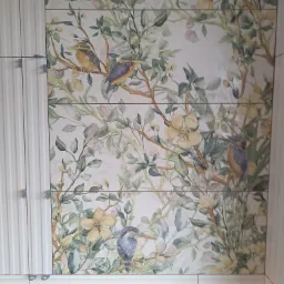 Ściana wyłożona płytkami ceramicznymi z motywem akwarelowych ptaków i kwitnących gałęzi, uzupełniona białymi szafkami z frezowanymi frontami.