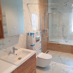 SAN-REMOnt - Nowoczesna łazienka z kabiną prysznicową, umywalką z drewnianą szafką i toaletą. Marmurowe płytki na ścianach i podłodze. Część elementów zabezpieczona taśmą.