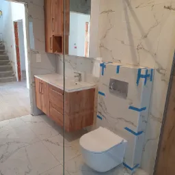 Nowoczesna łazienka z drewnianą szafką pod umywalką, marmurową płytką i podwieszaną toaletą z niebieską taśmą zabezpieczającą. Widok na niedokończone schody.