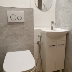 SAN-REMOnt - Nowoczesna łazienka po remoncie: biała umywalka z szafką, toaleta z przyciskiem Geberit, bidetka i lustro na tle szarych płytek imitujących marmur.