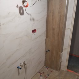 SAN-REMOnt - Łazienka w trakcie prac: białe płytki imitujące marmur na ścianach, drewnopodobne płytki na podłodze, widoczne instalacje i akcesoria montażowe.