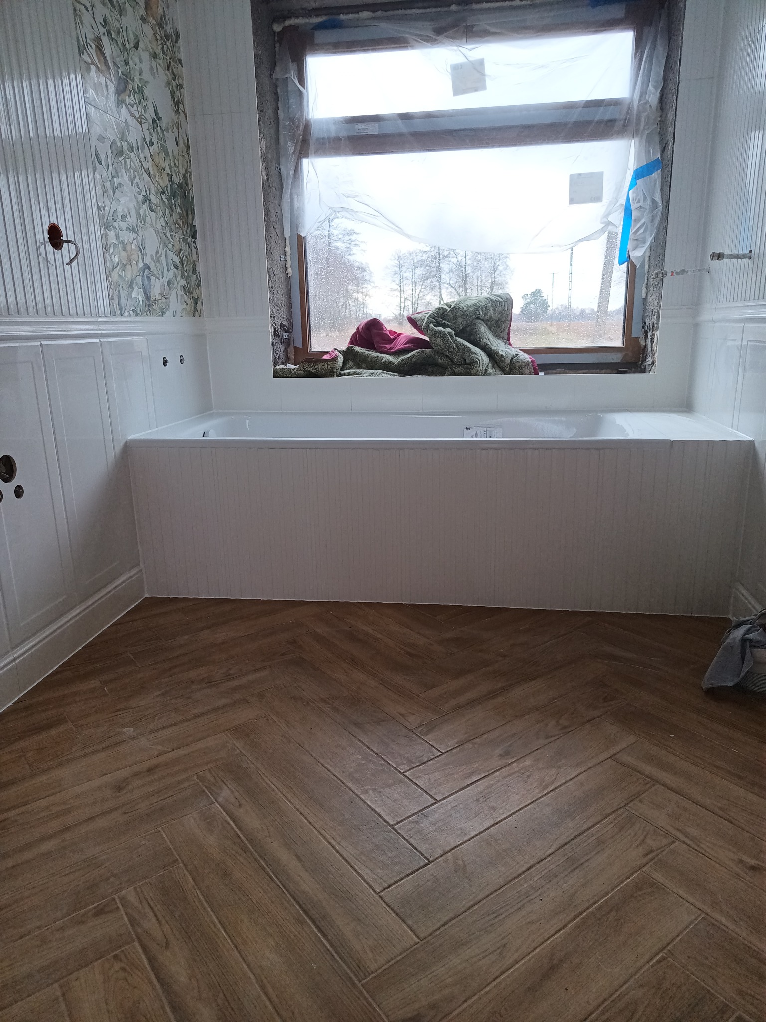 Wnętrze łazienki w trakcie prac wykończeniowych: ułożona podłoga z płytek imitujących drewno w jodełkę, obudowana wanna z pionowymi panelami, fragment ściany z tapetą w kwiaty, okno zabezpieczone...