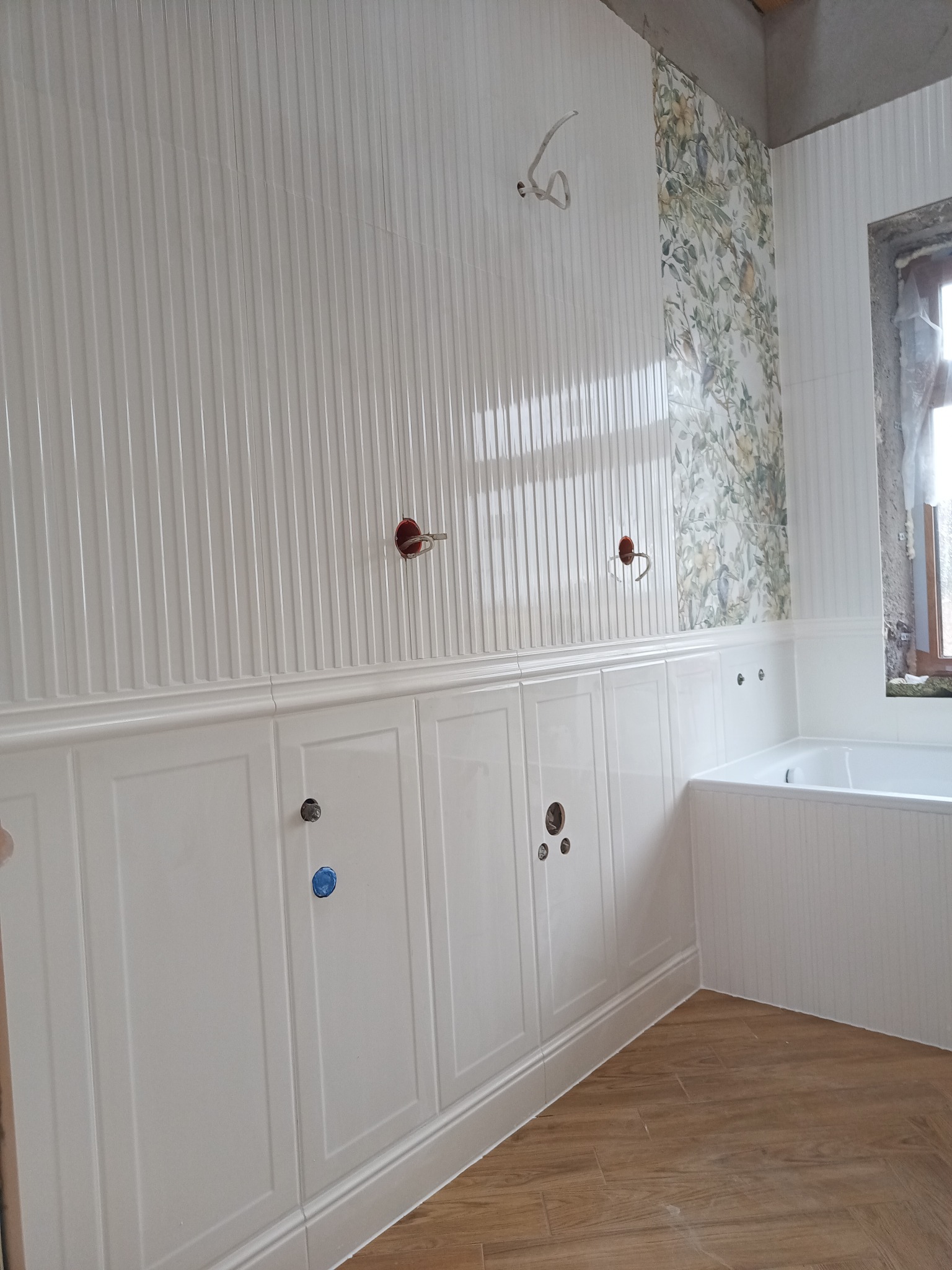 Wnętrze łazienki w trakcie prac wykończeniowych: białe płytki ścienne z pionowymi żłobieniami i panelami dolnymi, fragment ściany z motywem kwiatowym, przygotowane otwory na armaturę, widoczna...